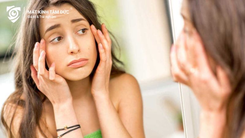 Người phụ nữ nhìn vào gương với đôi mắt sưng húp sau khi khóc, đang lo lắng về tình trạng của mắt.