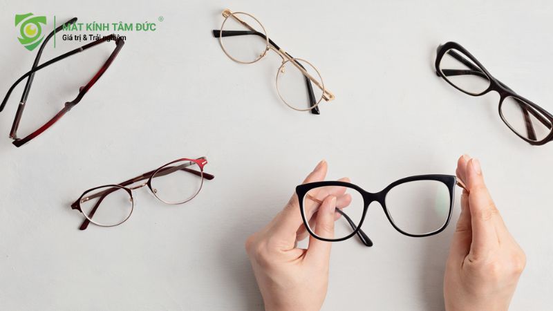 Hình ảnh một số cặp kính mắt khác nhau, bao gồm kính tròn, kính oval và kính vuông, được đặt trên bàn với bàn tay đang cầm một cặp kính.